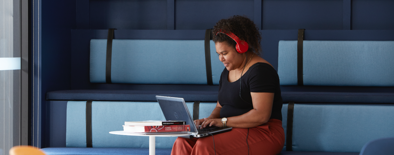 A young Indigenous student wearing headphones sits at a table, working on her laptop. She is smiling.