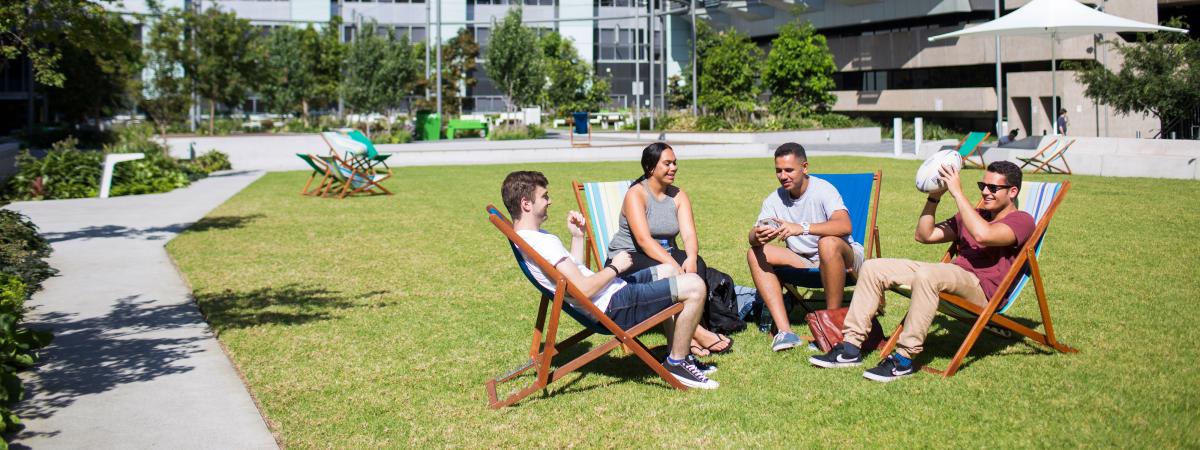 Four Indigenous students, three male and one female, sit on deck chairs on CUA Alumni Green, chatting and laughing. One is holding a football about to throw it back and forth with another male student. CUA buildings are in the background, including one showing the CUA sign.