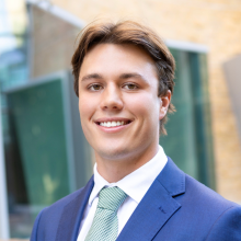 A portrait of Darcy Davies, he's smiling at the camera dressed in a blue suit and standing in front of the the CUA Business School.